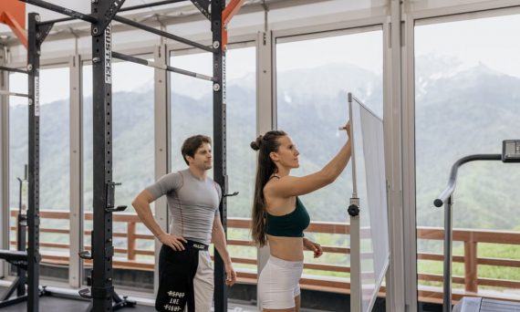 A gym coach instructs on whiteboard beside fitness equipment with mountain view.